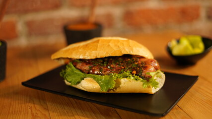 Choripan con chimichurri servido en un plato negro acompañado de salsa roja y verde, guacamole  y limones