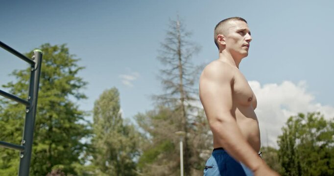 Muscular man walks away from the outdoor workout equipment in a park. He is shirtless and wearing blue shorts. He is getting ripped, shredded, and jacked at the outdoor gym.