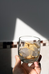 A glass filled with ice cubes and powdered Amanita muscaria mushroom on its surface, held in the hand, illuminated by natural sunlight. Preparing a homemade fly agaric elixir