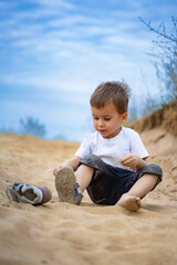 A young child playing in the sand on a sunny day. A boy sits on a sandy path, focused on putting on his shoe, enjoying a sunny day outdoors with a clear blue sky above.