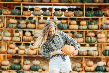 young woman stands at pumpkin festival, holding large orange and green pumpkin. She is surrounded by rows of pumpkins and a wooden display. The autumn farm setting captures the harvest season vibe.