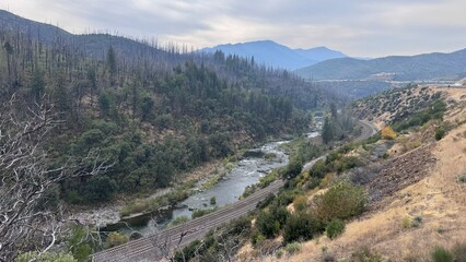 The railway runs between mountains and hills along the bottom of a ravine along the river in Oregon. Against the background of the mountain, a partially burned pine forest can be seen in the haze