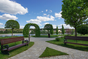 A well-maintained park with a beautiful landscape against a blue sky with white clouds. Wooden benches, tiled paths.