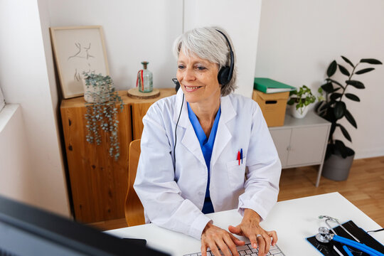 Senior doctor in white coat and headset working on laptop at office desk. Professional practitioner woman typing on keyboard looking at computer screen at clinic workplace. Healthcare people concept. - Powered by Adobe