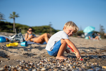 Young Boy Playing with Shovel on Pebble Beach While Mother Relaxes in the Background