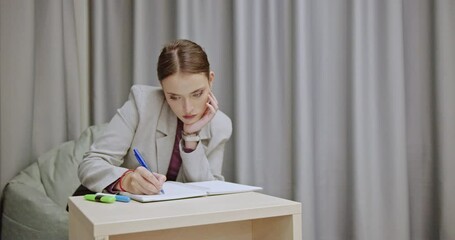 Young female student in stylish formal wear studying in modern library or home, sitting on a comfy beanbag. She writes in her notebook and uses colorful highlighters.