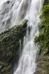 Fototapeta premium The image showcases a spectacular waterfall cascading over rugged, moss-covered rocks, surrounded by rich greenery, creating a serene yet powerful atmosphere of natural beauty.