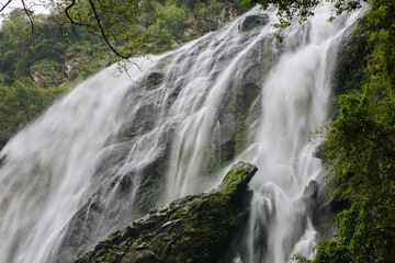 Obraz premium The image showcases a stunning waterfall flowing over jagged rocks, surrounded by vibrant vegetation. The atmosphere evokes tranquility with a hint of mist in the air, enhancing nature's artistry.