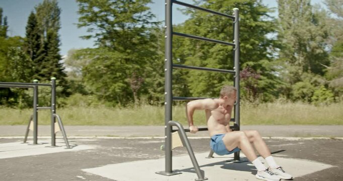 A muscular man working out at an outdoor gym in a park, using the parallel bars to do dips. He is shirtless and focused on his fitness.