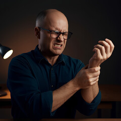Naklejka premium A man grimacing in pain while holding his wrist, possibly due to strain or injury, seated indoors.