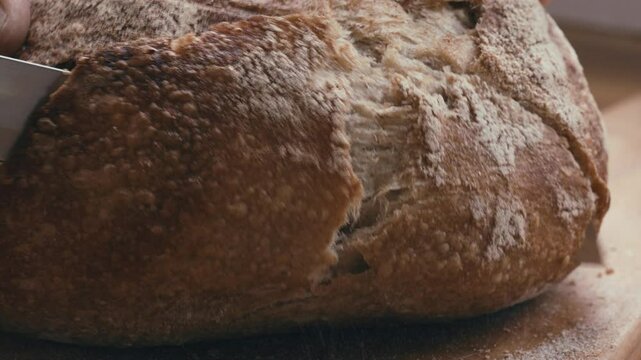 Hand slicing freshly baked loaf of crusty bread with serrated knife. Extreme close-up view