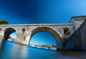 Fototapeta premium An arched stone bridge spans a blue body of water, with a clear blue sky above.