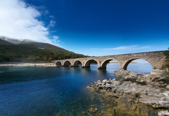 Fototapeta premium Stone arch bridge spans a calm, clear blue body of water, with a green hillside beyond.