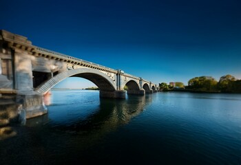 Fototapeta premium A stone bridge with multiple arches spans a calm river, with a blue sky above and greenery along the banks.