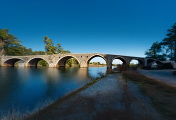 Fototapeta premium A stone bridge arches over a calm river, surrounded by greenery under a blue sky.