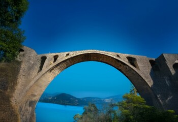 Fototapeta premium A stone arch bridge spans a body of water, with a clear blue sky and hills in the background.