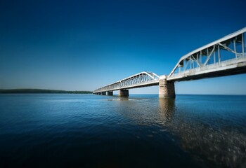 Fototapeta premium A metal bridge spans a wide body of water under a clear blue sky.