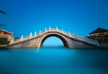 Obraz premium A white stone bridge arches over a still blue body of water, a clear blue sky above.