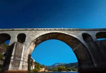 Fototapeta premium A stone arch bridge curves against a bright blue sky, with a glimpse of green trees and hills in the background.