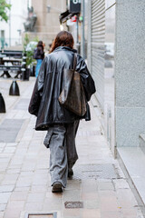 A woman dressed in a stylish black leather jacket is leisurely walking down a bustling sidewalk on a sunny day in the city