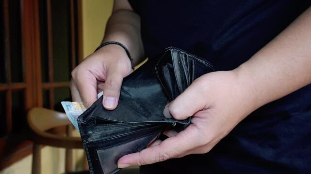 A close-up of a man's hand realizing that his wallet is empty only contains one 2.000 Indonesian Rupiah note
