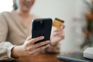 A young woman is sitting at her desk, smiling as she holds her phone and a credit card, likely making an online purchase. She is in a modern, bright office environment