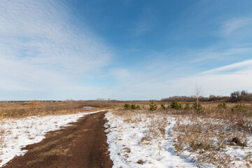 Dirt road in a field overgrown with grass. Autumn landscape. The first snow is melting. There is a blue autumn sky above the horizon.