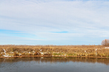 Natural lake in the countryside, the water is covered with the first ice, which reflects the blue sky. The shore is covered with dry grass. Autumn, sunny, frosty day.