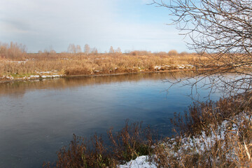 The first ice of autumn. Autumn landscape with melting snow on the shore with dry grass in early winter. The surface of the lake is covered with thin ice. Sunny, frosty day.