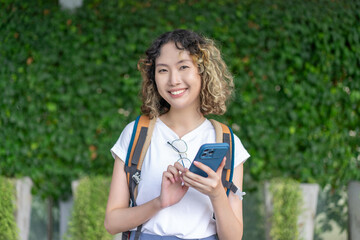 Asian cheerful young woman holding a smartphone, standing outdoors with a smile. Perfect for travel and lifestyle concepts.