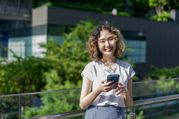 Asian cheerful young woman holding a smartphone, standing outdoors with a smile. Perfect for travel and lifestyle concepts.
