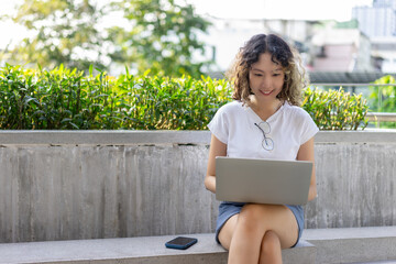 Asian young woman sits outdoors using a laptop, smiling while working in an urban setting. Perfect for remote work, technology, and lifestyle themes.