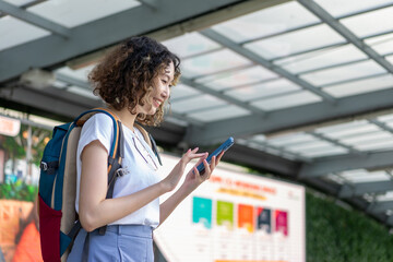 Asian cheerful young woman holding a smartphone, standing outdoors with a smile. Perfect for travel and lifestyle concepts.