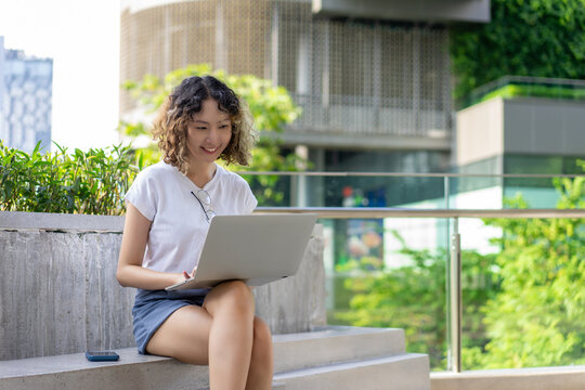 Asian young woman sits outdoors using a laptop, smiling while working in an urban setting. Perfect for remote work, technology, and lifestyle themes.