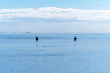 Silhouettes of two people fishing in calm waters standing waist deep under a blue sky in the calm Mediterranean Sea at the mouth of the Ebro River, with Trabucador beach on the horizon.
