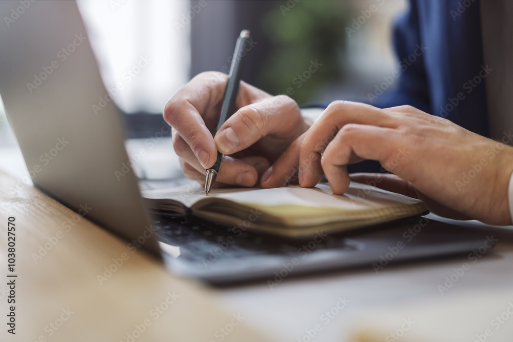 Poster detailed view of a man's hand jotting down notes in a notepad on a modern laptop, with a blurred bac
