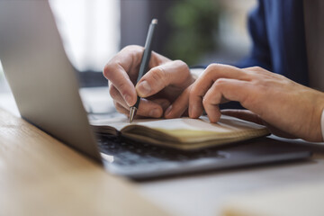 Detailed view of a man's hand jotting down notes in a notepad on a modern laptop, with a blurred...