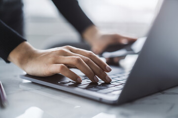 A close view of a woman's hands typing on a modern laptop keyboard, office surroundings appearing fuzzy in the backdrop