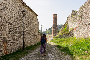 Tourist woman walking in charming cobblestone alleyway towards ancient red mosque in Berat castle, Albania. Weathered stone walls partially collapsed creating sense of history and decay. Xhamia e Kuqe