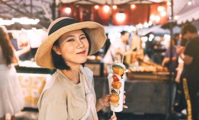 Happy young asian foodie woman eating bbq grilled skewers at outdoor night market street food vendor