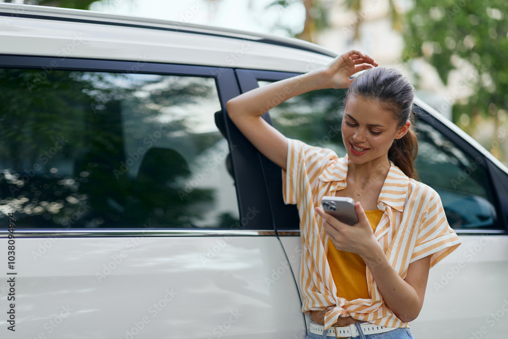 Wall mural Young woman using smartphone while leaning against a white car, smiling and enjoying communication, wearing a stylish outfit with striped shirt and casual jeans, outdoor background