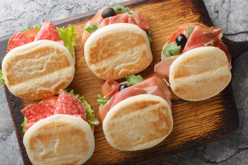 Small italian tigelle sandwiches with sausage, jamon and fresh vegetables close-up on wooden board on table. Horizontal top view from above