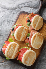 Popular street food in Emilia-Romagna, tigelle or crescentine sandwich with salami, ham and fresh vegetables close-up on wooden board on table. Vertical top view from above