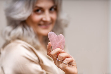 Jade Gua Sha Scraper. Portrait of a woman holding a Gua Sha jade scraper in her hand, isolated on a beige background with space for text. Concept of skin care and keeping youthfulness