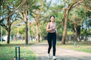 Active young Asian sports woman running in green park outdoors with sunlight in morning. Health and fitness training, healthy living lifestyle, sports routine concept