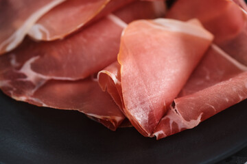 Thin italian prosciutto slices on black plate on conctere background closeup