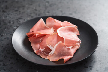 Thin italian prosciutto slices on black plate on conctere background