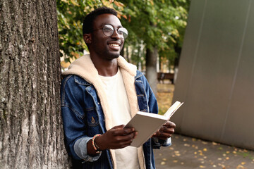 Portrait of happy smiling young african man student reading a book standing in autumn city park