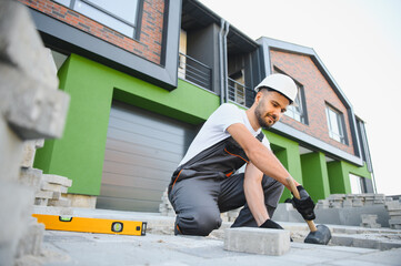 The master in gloves lays paving stones in layers. Garden brick pathway paving by professional paver worker. Laying gray concrete paving slabs in house courtyard on sand foundation base