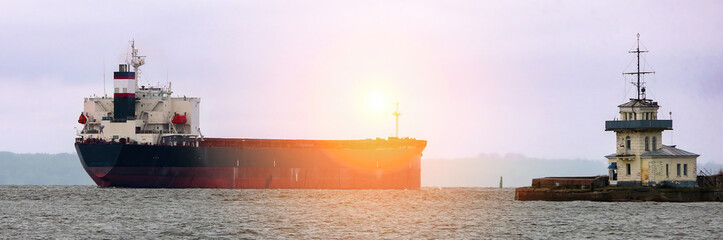 Fototapeta premium A cargo ship navigates through coastal waters near a lighthouse under a twilight sky